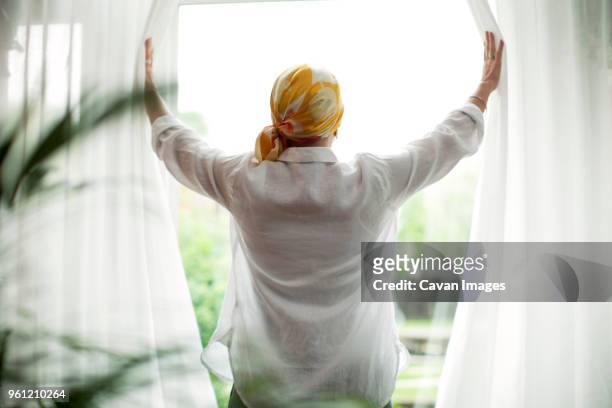 rear view of mature woman opening curtains at window - sick window stockfoto's en -beelden