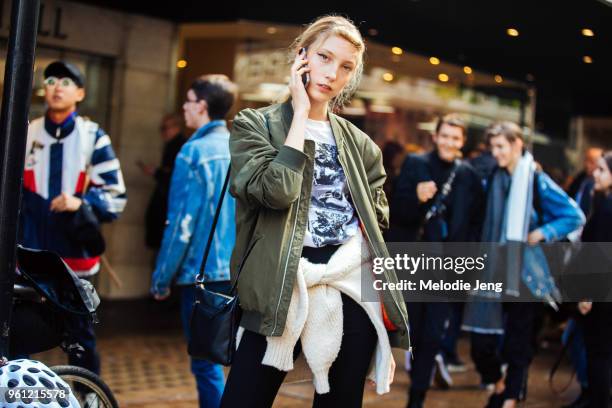 Model Sasha Komarova talks on the phone and wears an olive bomber jacket, white graphic shirt, sweater around her waist, and black purse during...