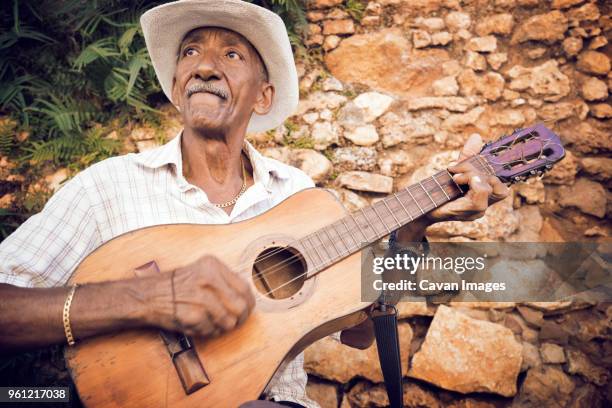 man playing guitar while sitting against stone wall - plucking an instrument - fotografias e filmes do acervo