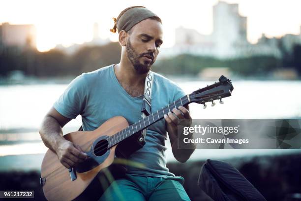 man playing guitar while sitting at lakeshore during sunset - acoustic guitarist stock pictures, royalty-free photos & images