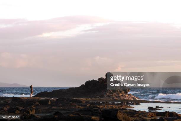 distant view of woman standing on rock by sea against cloudy sky - kona coast stock pictures, royalty-free photos & images