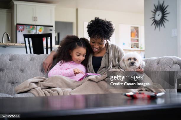 mother looking at daughter using tablet computer while sitting with lhasa apso on sofa at home - girl on couch with dog foto e immagini stock
