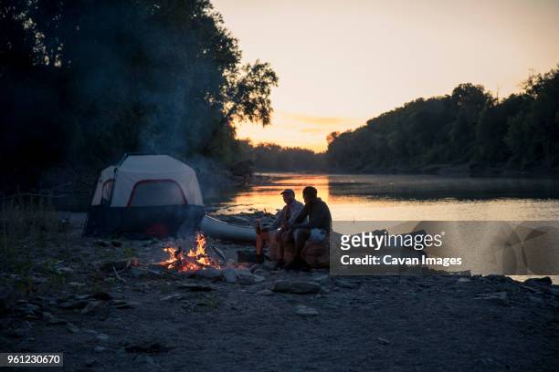 male friends relaxing at campsite by lake against sky during sunset - male friendship stock pictures, royalty-free photos & images