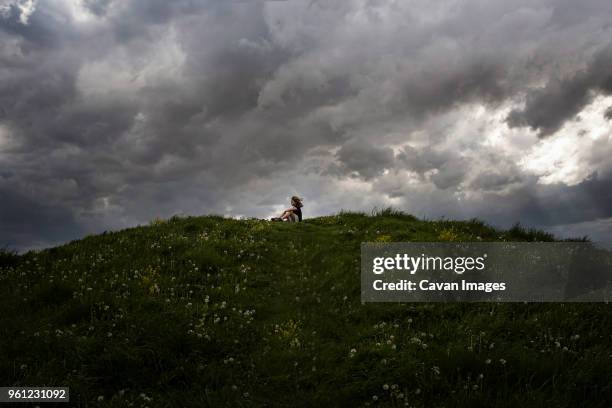 low angle view of girl sitting on hill against stormy clouds - girl mound stock pictures, royalty-free photos & images