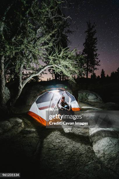 hiker sitting in illuminated tent on rock by trees at night - big bear lake stockfoto's en -beelden