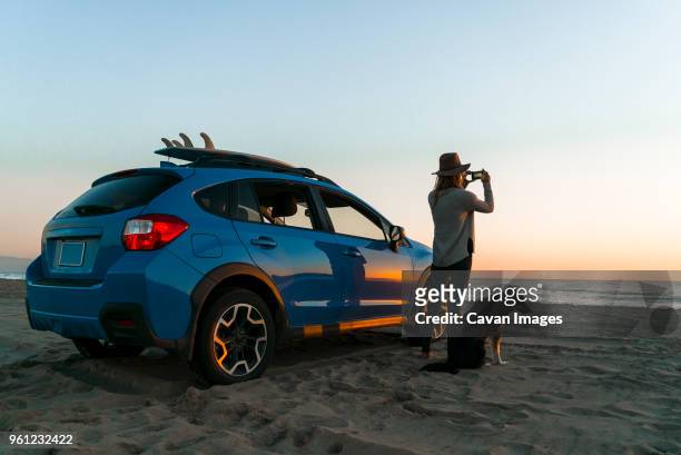 woman photographing while standing with dog by car at beach - véhicule terrestre photos et images de collection