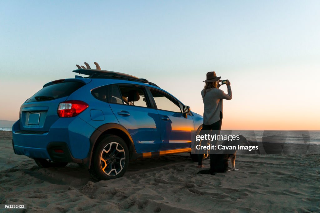 Woman photographing while standing with dog by car at beach