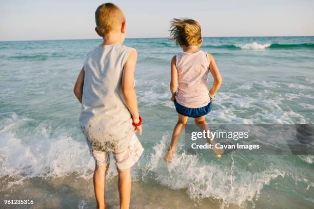 rear view of siblings playing with waves at beach - destin stock pictures, royalty-free photos & images