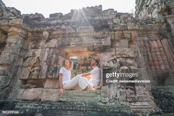 young couple contemplating ancient temple, sitting on window in stone wall looking around and relaxing taking the time to breathe it all in, sunset time sunbeam - angkor wat stock pictures, royalty-free photos & images