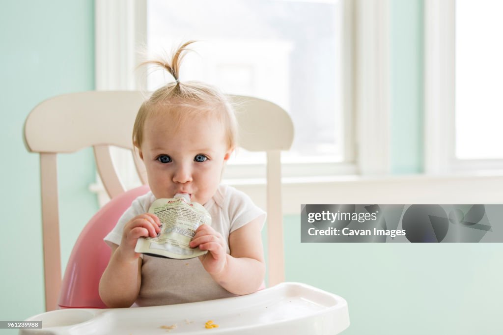 Portrait of cute baby girl eating apple sauce from pack while sitting on high chair at home