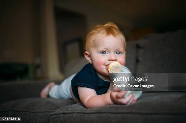 portrait of cute baby boy drinking milk from bottle while lying on sofa at home - mjölkflaska bildbanksfoton och bilder