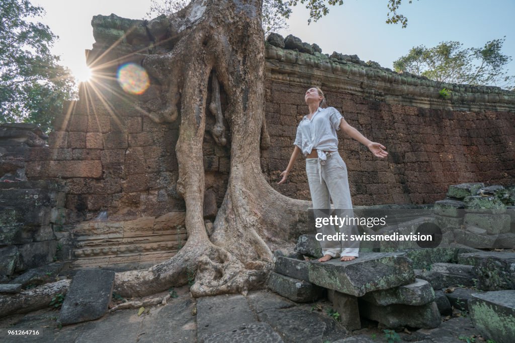 Young woman exercising yoga in ancient temple near majestic tree roots; sunlight passing through and relaxing with some meditation; concept of people with healthy lifestyles and wellbeing and travel