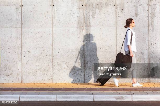 joven mujer caminando en una acera al lado de la pared de hormigón y tirando una pequeñas maletas con ruedas - maleta fotografías e imágenes de stock