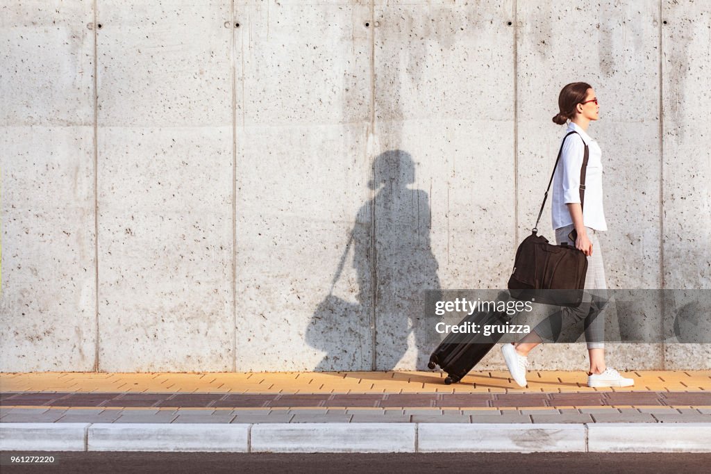 Jovem mulher caminhando na calçada, ao lado da parede de concreto e puxando uma mala com rodas pequena