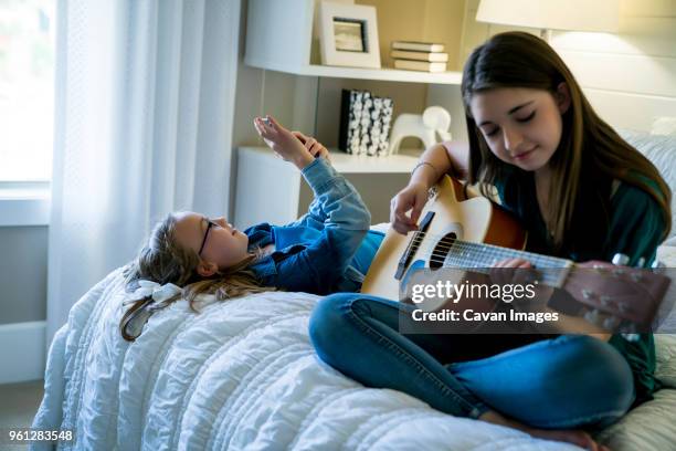 girl using smart phone while sister playing guitar on bed - plucking an instrument - fotografias e filmes do acervo