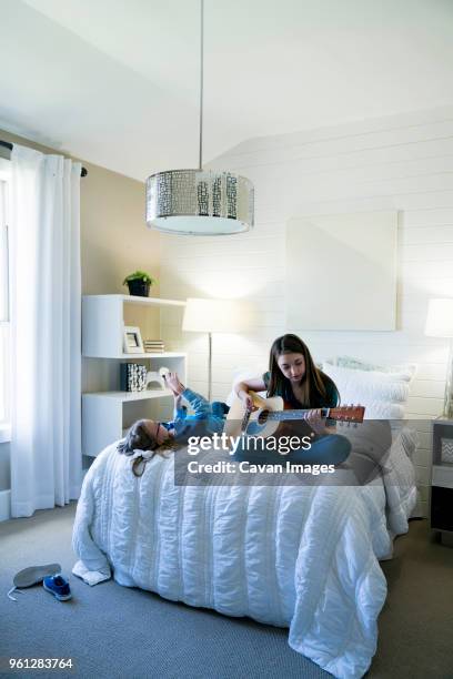 girl using smart phone while sister playing guitar on bed in bedroom - plucking an instrument - fotografias e filmes do acervo