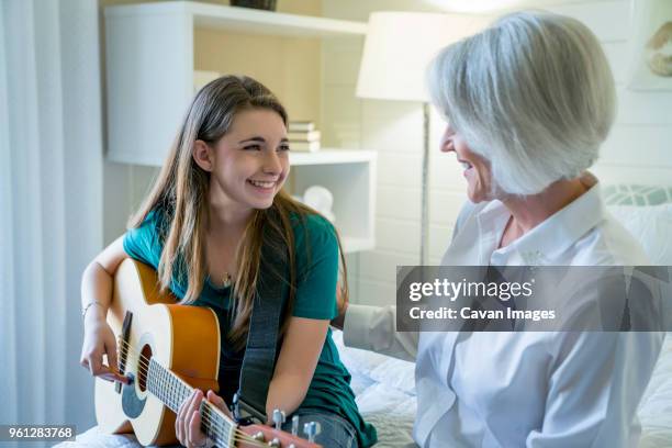 happy granddaughter playing guitar while sitting by grandmother in bedroom - plucking an instrument - fotografias e filmes do acervo