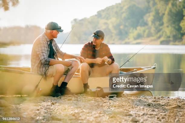 friends talking while sitting by fishing rods on boat at lakeshore - fishing rod stock-fotos und bilder