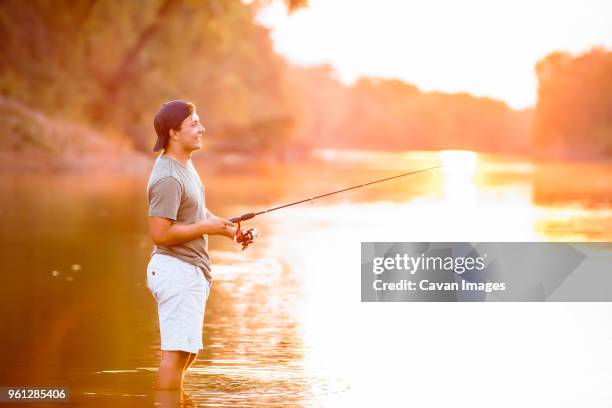 side view of young man fishing while standing in lake during sunset - young men fishing stock pictures, royalty-free photos & images