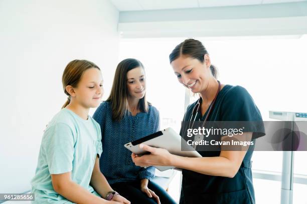 cheerful pediatrician showing tablet computer to mother and daughter sitting on examination table in hospital - medical examination room stock pictures, royalty-free photos & images