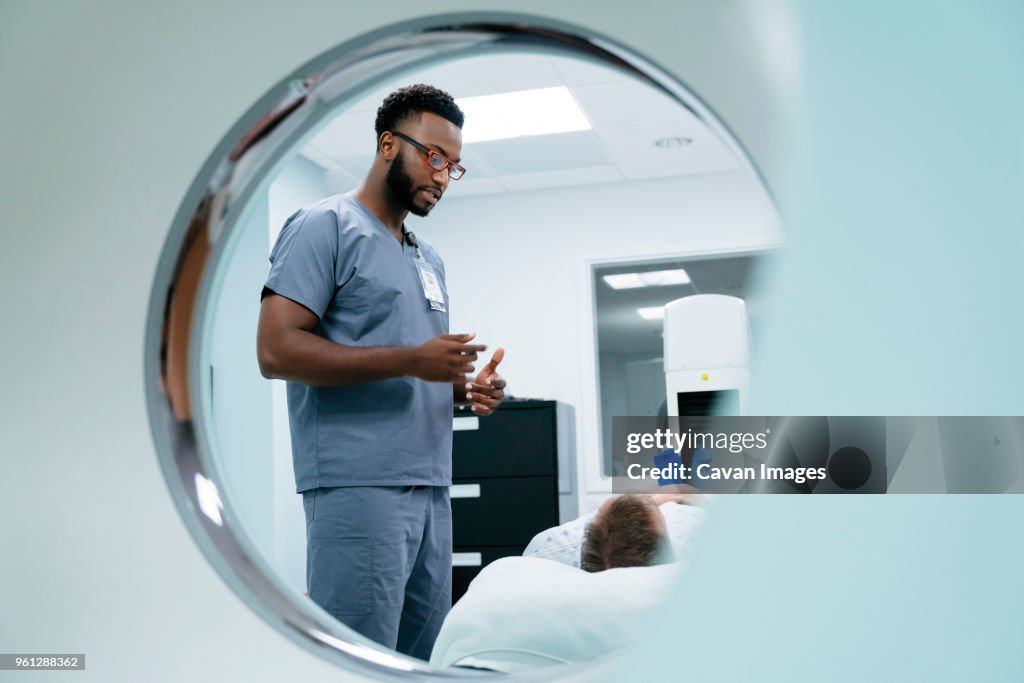 Male nurse talking with patient lying in examination room seen through MRI Scanner