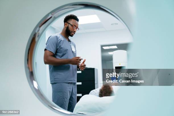 male nurse talking with patient lying in examination room seen through mri scanner - hospital gown stock photos et images de collection