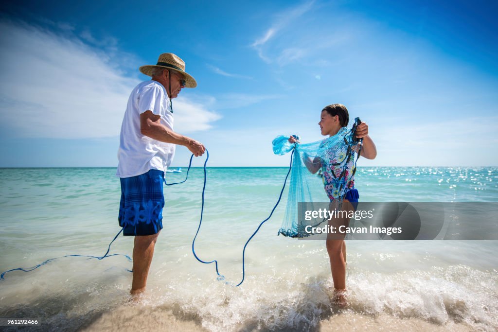 Grandfather holding rope of fishing net carried by granddaughter while standing on shore at beach against sky