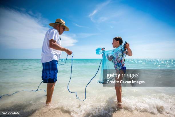 grandfather holding rope of fishing net carried by granddaughter while standing on shore at beach against sky - tampa stock pictures, royalty-free photos & images