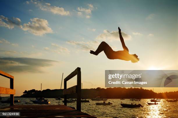 silhouette man backflipping into sea against sky during sunset - salto de espalda fotografías e imágenes de stock