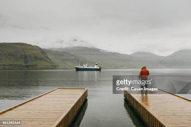 rear view of man in hooded jacket standing on pier against mountains during foggy weather - îles féroé photos et images de collection