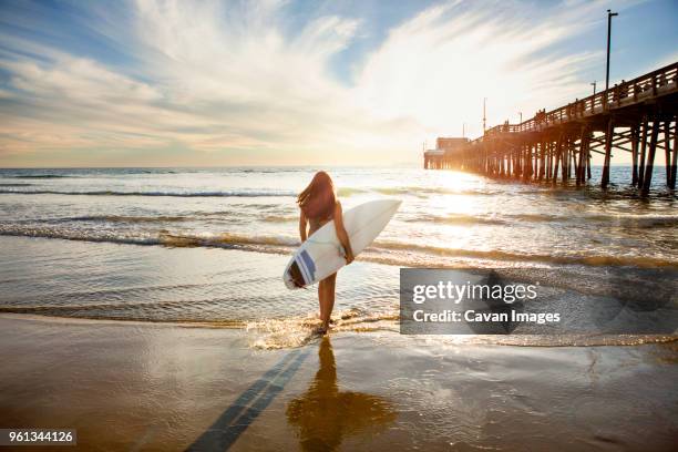 rear view of woman holding surfboard and standing on shore - newport beach stock-fotos und bilder