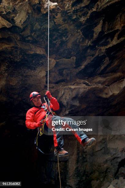 full length of man hanging on rope by rock formation in cave - espeleología fotografías e imágenes de stock