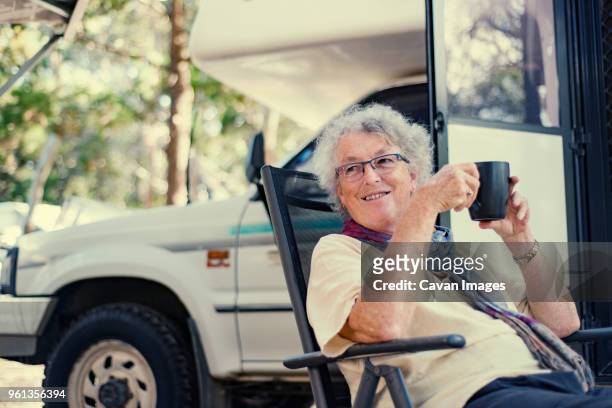 happy senior woman holding coffee cup while sitting by travel trailer in forest - australian cup day stock pictures, royalty-free photos & images