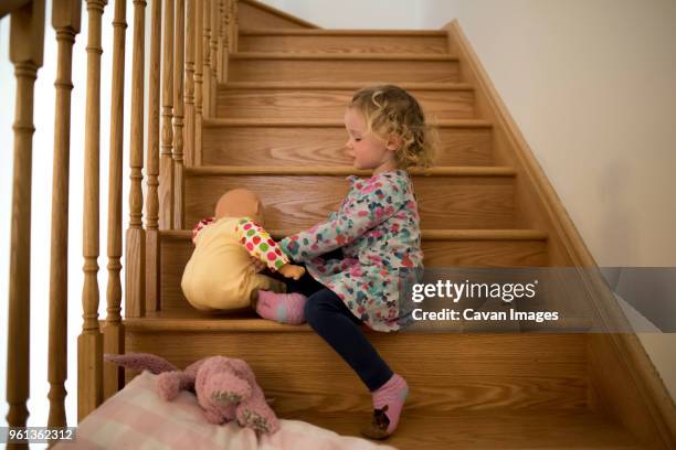 side view of girl playing with doll while sitting on wooden steps at home - doll house stockfoto's en -beelden