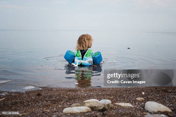 rear view of girl wearing life jacket and water wings while sitting in lake simcoe against sky - life jacket isolated stock pictures, royalty-free photos & images