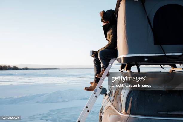 side view of man holding cup while sitting in roof tent on car against sky - car roof tent stock pictures, royalty-free photos & images