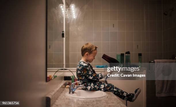 side view of boy holding hair dryer while sitting by bathroom sink at home - föhn stock-fotos und bilder