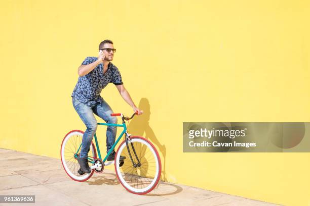 full length of man talking on smart phone while riding bicycle against yellow wall at sidewalk in city during sunny day - guy with phone full image ストックフォトと画像