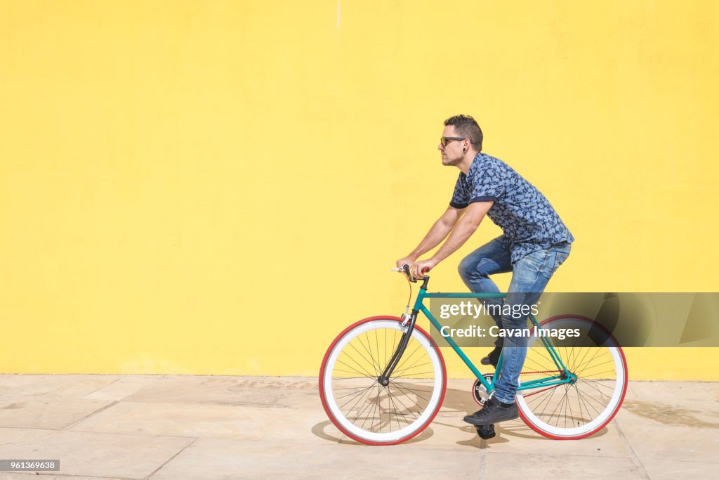 Full length of man riding bicycle against yellow wall at sidewalk in city