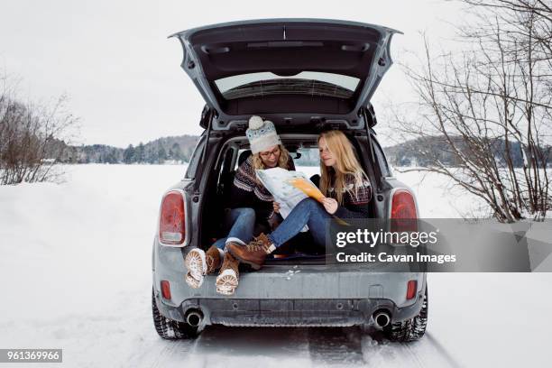 happy female friends reading map while sitting in car trunk during winter - winter road trip stock pictures, royalty-free photos & images