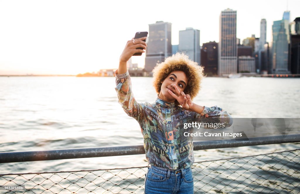 Woman taking selfie through mobile phone while standing against river