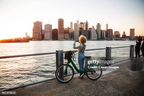 woman standing by bicycle photographing river and city - new york trip stock pictures, royalty-free photos & images