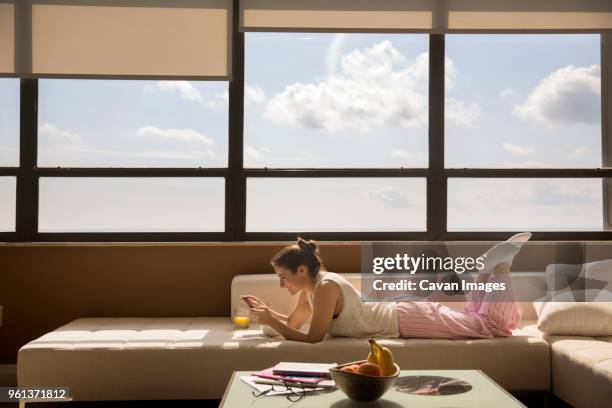 happy woman using smart phone while eating breakfast on sofa at home - allongé sur le devant photos et images de collection