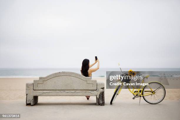 woman taking selfie while sitting on bench at beach against clear sky - manhattan beach stock pictures, royalty-free photos & images