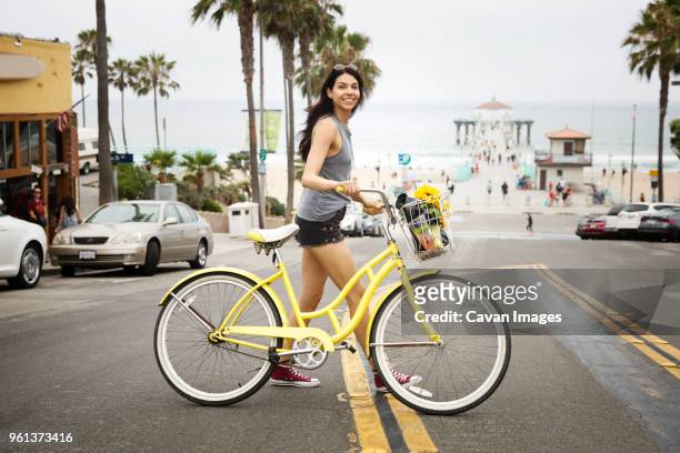 side view of smiling woman with bicycle crossing city street - manhattan beach stock-fotos und bilder