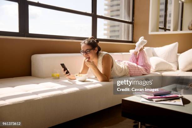 woman using smart phone while eating breakfast on sofa at home - juice concentrate stock pictures, royalty-free photos & images