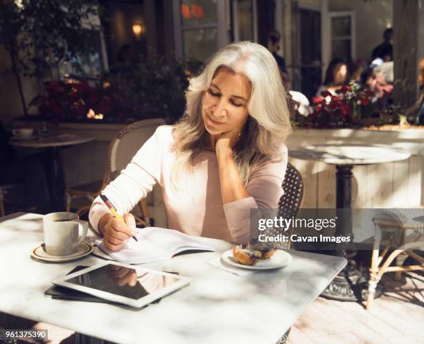 mature woman writing in book at restaurant table - 50s woman writing at table imagens e fotografias de stock