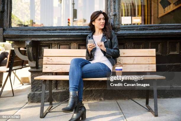 thoughtful woman holding smart phone while sitting on bench at sidewalk cafe - new york cafe stock-fotos und bilder