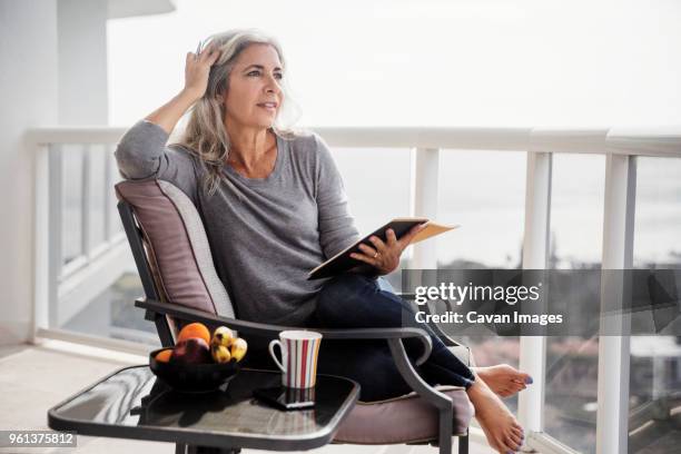 thoughtful woman holding book while sitting on chair on balcony - tisch betrachten stock-fotos und bilder