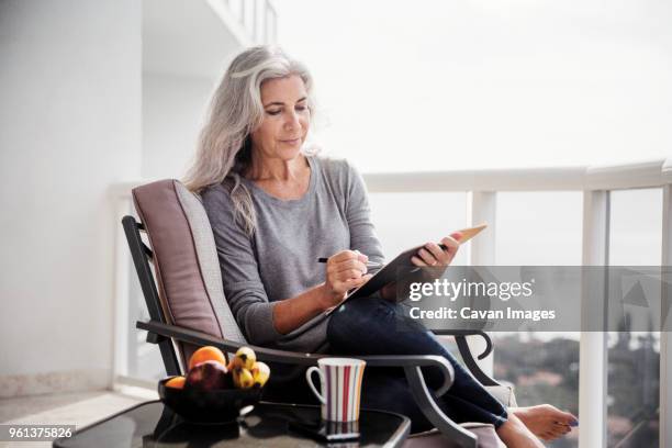 mature woman writing in book while sitting on chair on balcony - 50s woman writing at table imagens e fotografias de stock